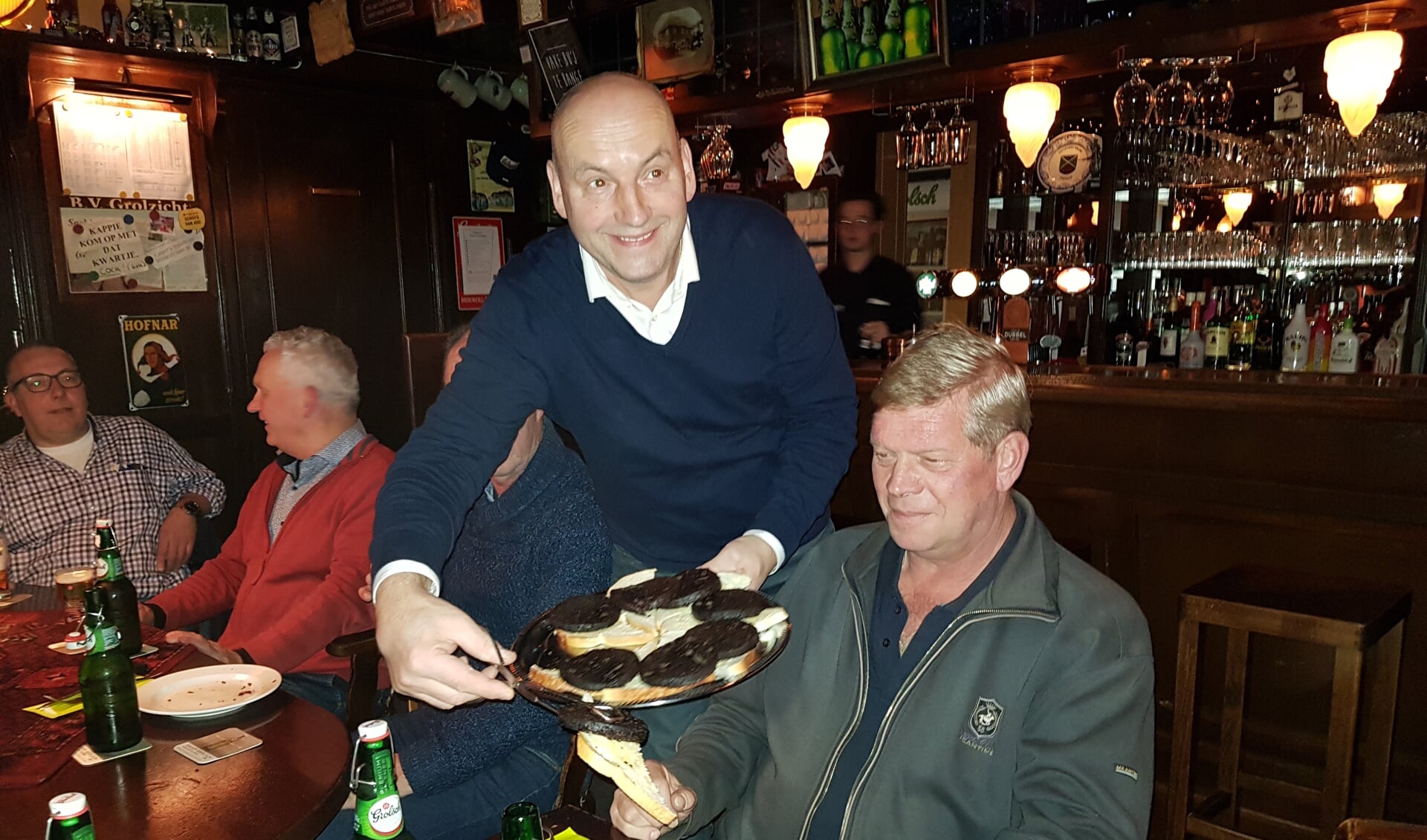 Casper van Uem serveert Rudi Spekschoor de bakbloedworst op een sneetje witbrood. Geheel links Ronnie Wopereis.