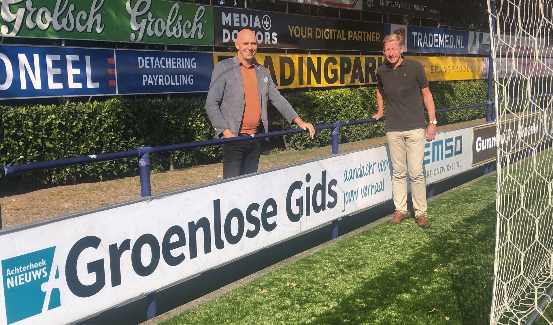 Patrick Verheij van Achterhoek Nieuws (links) en Erik Schuurmans van CT Grol bij het reclamebord van Groenlose Gids op het hoofdveld van Sportpark Den Elshof. Foto: PR Grol