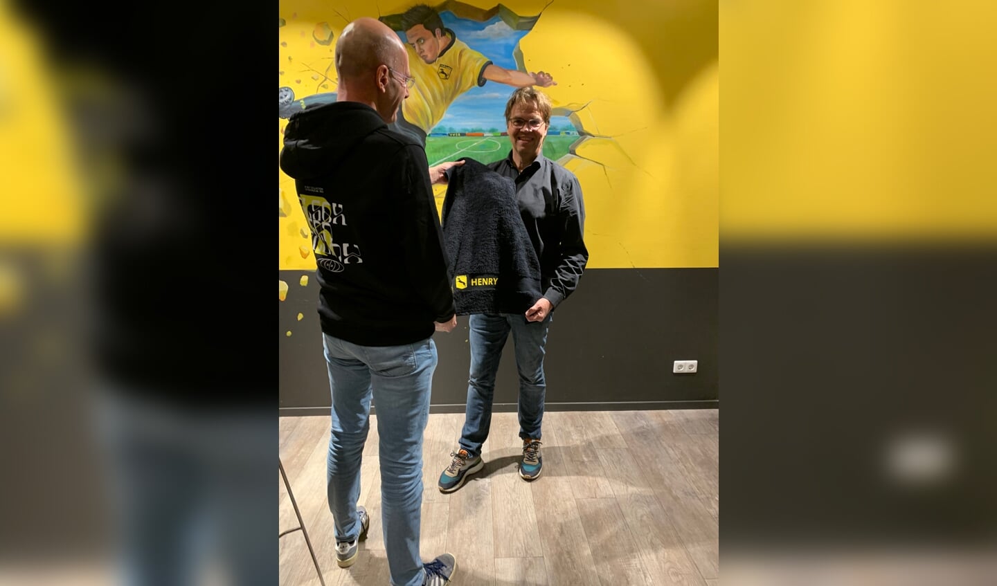 Voorzitter Henry Meutstege ontvangt de handdoek met zijn naam uit handen van een lid van de jubileumcommissie gehuld in de Ruurlo-hoodie. Foto: PR