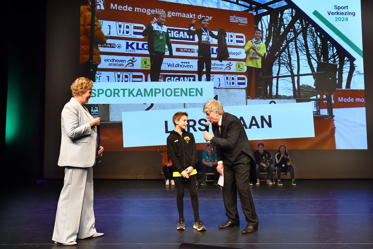 Wethouder Ria Ankersmit (links) kijkt toe hoe Lars Kraan (10) aan Frans Miggelbrink uitlegt hij hoe zijn bronzen plak veroverde op het NK cross.