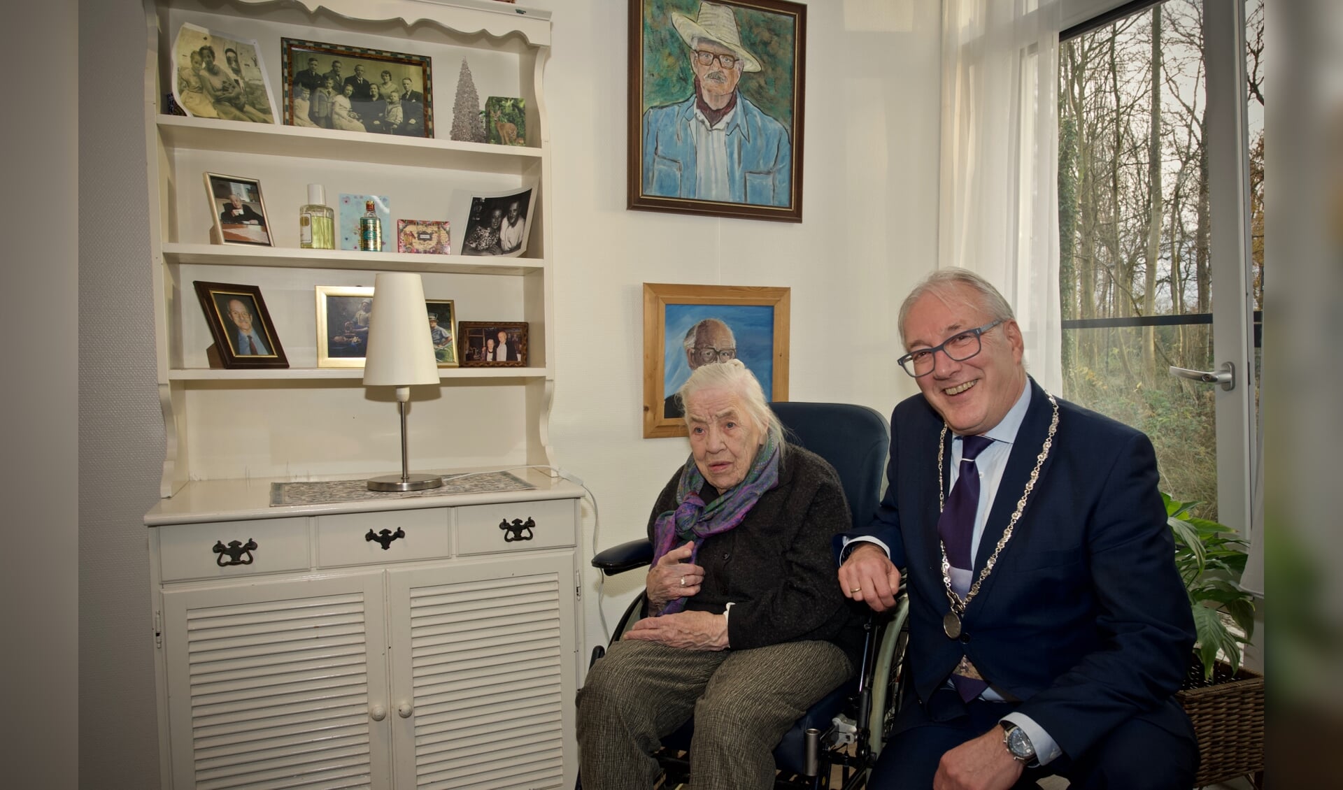 Mevrouw Hulsman samen met burgemeester Hans Romeyn bij een kast met de vele foto's en schilderijen geschilderd door meneer Hulsman.