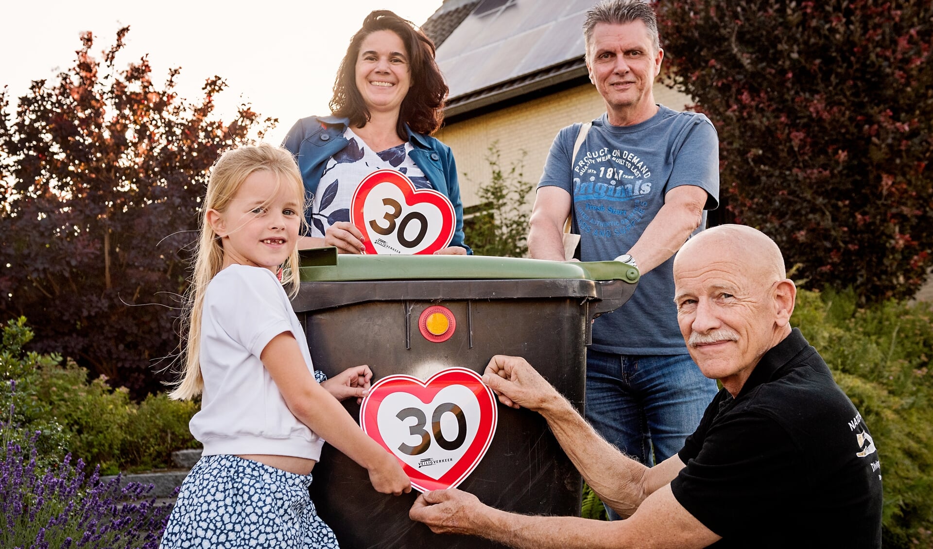 Jasmijn en opa Wolf plakken de 30 kilometer sticker van Veilig Verkeer Nederland, die Karina van de Goor en Jack Stevelmans net hebben uitgereikt, op de groen-container.