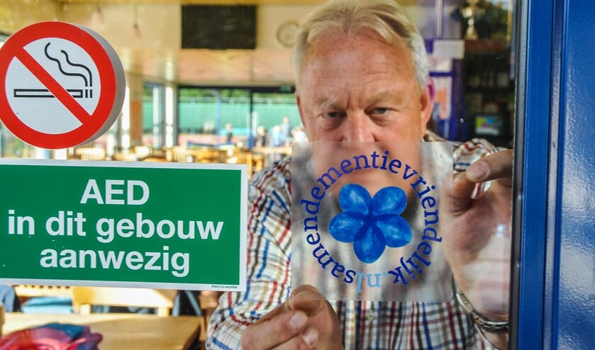 <p>WTC-voorzitter Cor Bavinck hangt de dementievriendelijke sticker op.</p>