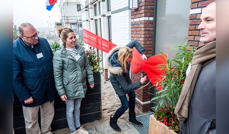 Lianka mag vlak voor haar voordeur het lintje van de eerste huizen doorknippen. Partner Martijn en buurvrouw Floor kijken samen met wethouder Vos toe.