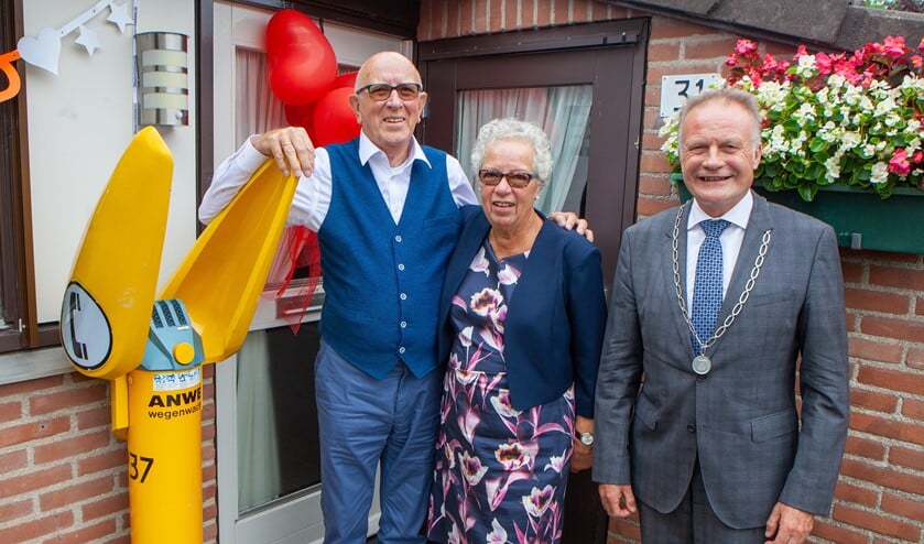 Jan en Renée met de burgemeester voor de deur van hun huis, waar de Wegenwachtpaal herinnert aan het werk van Jan bij de ANWB.