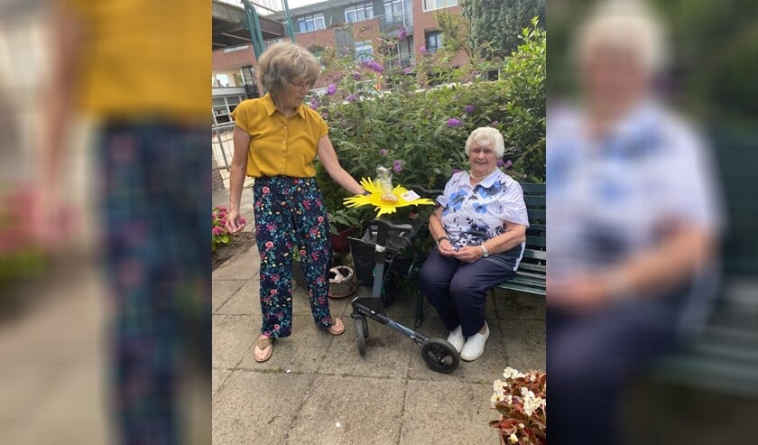 <p>Vrijwilliger Tineke Schouten overhandigt een taartje op zonnig bordje aan Zonnebloemgast Bep Post (92).</p>