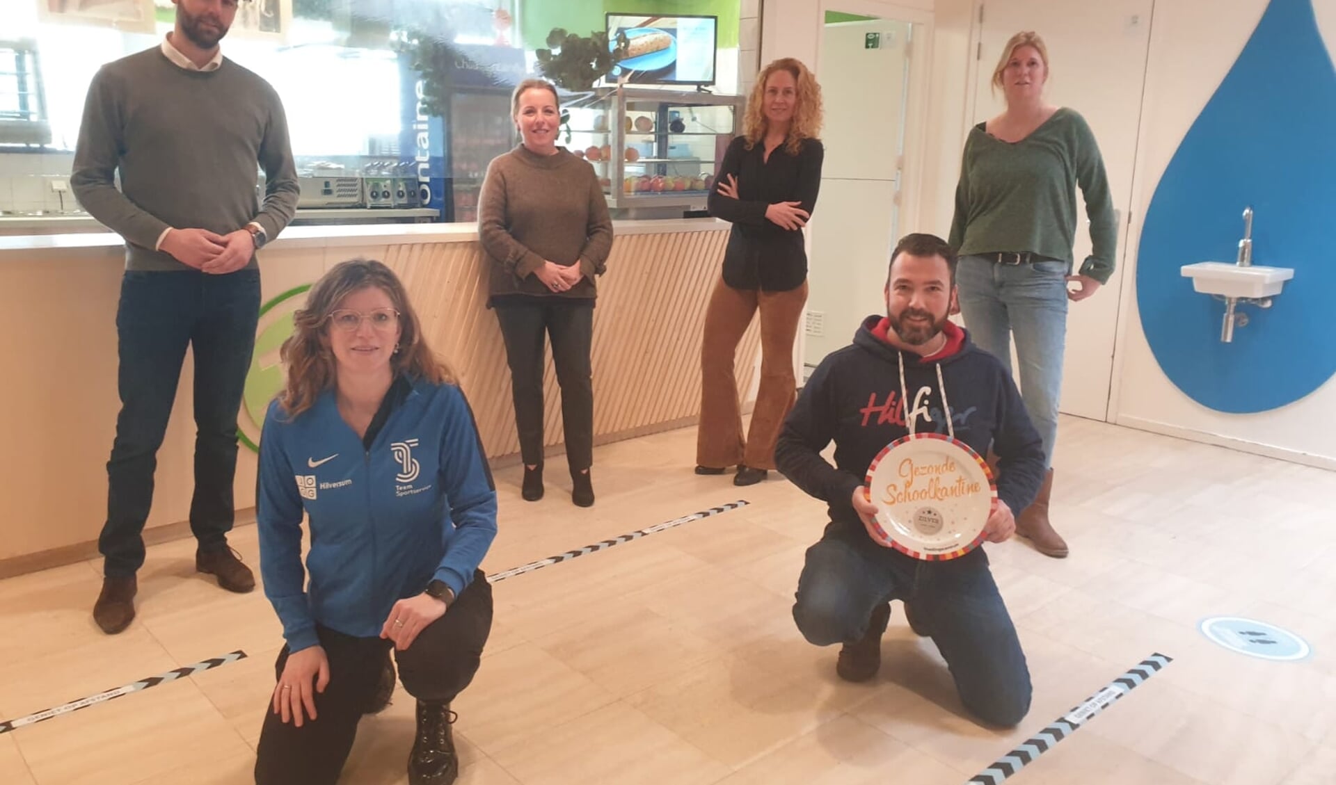 Van linksaf: Rector Robert van der Sijde, Femke Roest (JOGG-regisseur gemeente Hilversum), Carola van den Berg, buurtcoördinator, voedingsadviseur Mandy Evers, kantinebeheerder Rauko Verhoog en kantinemedewerker Hanneke Robeer poseren coronaproof. 