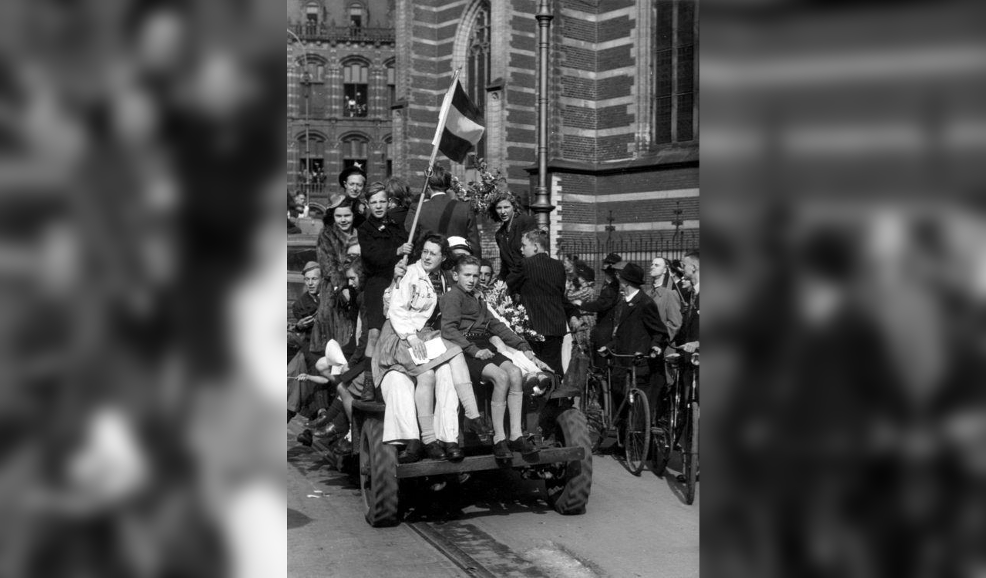 Een jeep vol met feestvierende mensen en een Nederlandse vlag rijdt langs de Nieuwe Kerk naar de Dam bij de bevrijding van Amsterdam aan het eind van de Tweede Wereldoorlog, Nederland 8 mei 1945.
