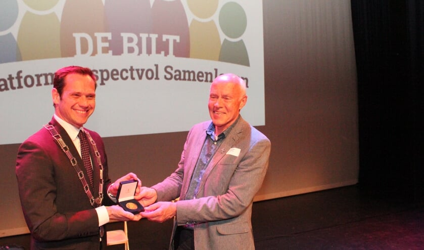 Burgemeester Sjoerd Potters overhandigt de Chapeaupenning aan Gert Landman, met op de achtergrond een deel van het nieuwe logo van het Platform Respectvol Samenleven.