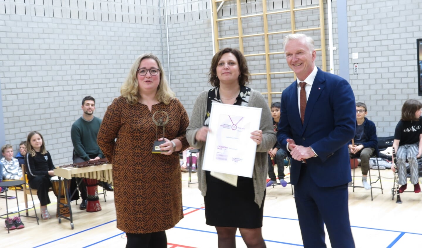 Nadine ten Kate en Linda Kuijt nemen trots de prijs voor het muziekonderwijsproject van de Julianaschool in ontvangst van gedeputeerde Robert Strijk.