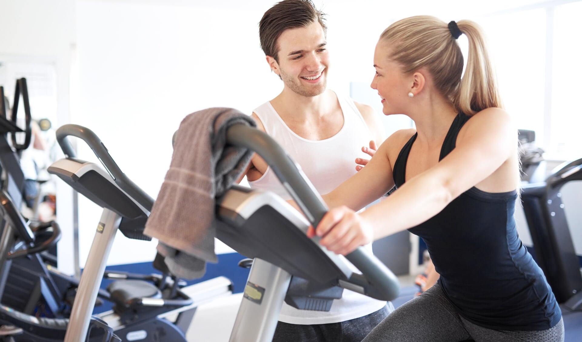 Handsome Young Fitness Trainer Explaining Something to a Young Woman While on Elliptical Bike Device Inside the Gym