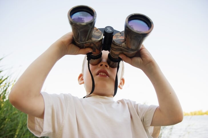 Portrait of little boy looking through binoculars on river bank. Cute kid with binoculars. The boy looks up at the sky through binoculars