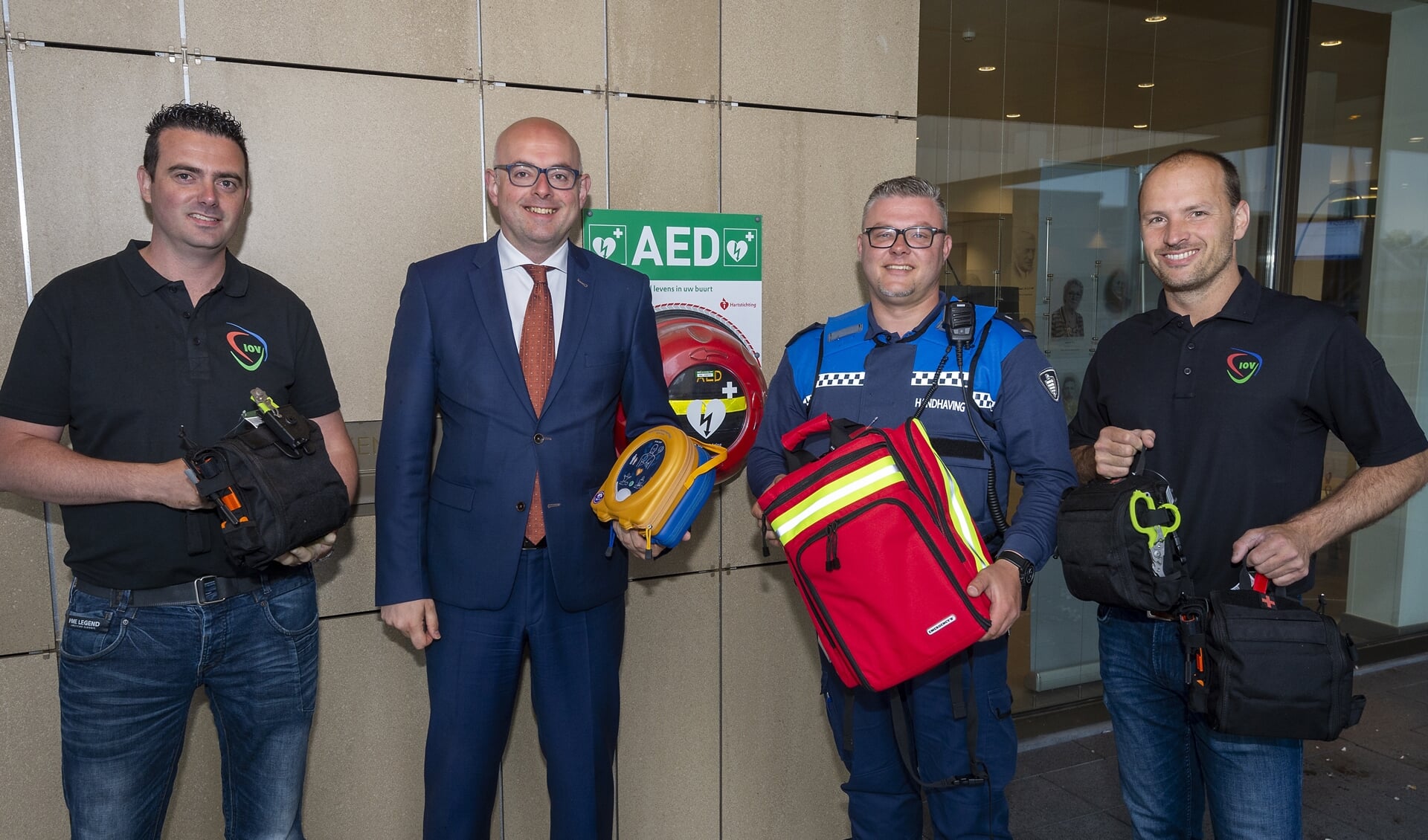 • V.l.n.r.: Marcel Beekman (IOV), Burgemeester Vroom, BOA Xander en Mark Schileroord (IOV). BOA Xander neemt de AED en trauma- en EHBO-pakket in ontvangst. 