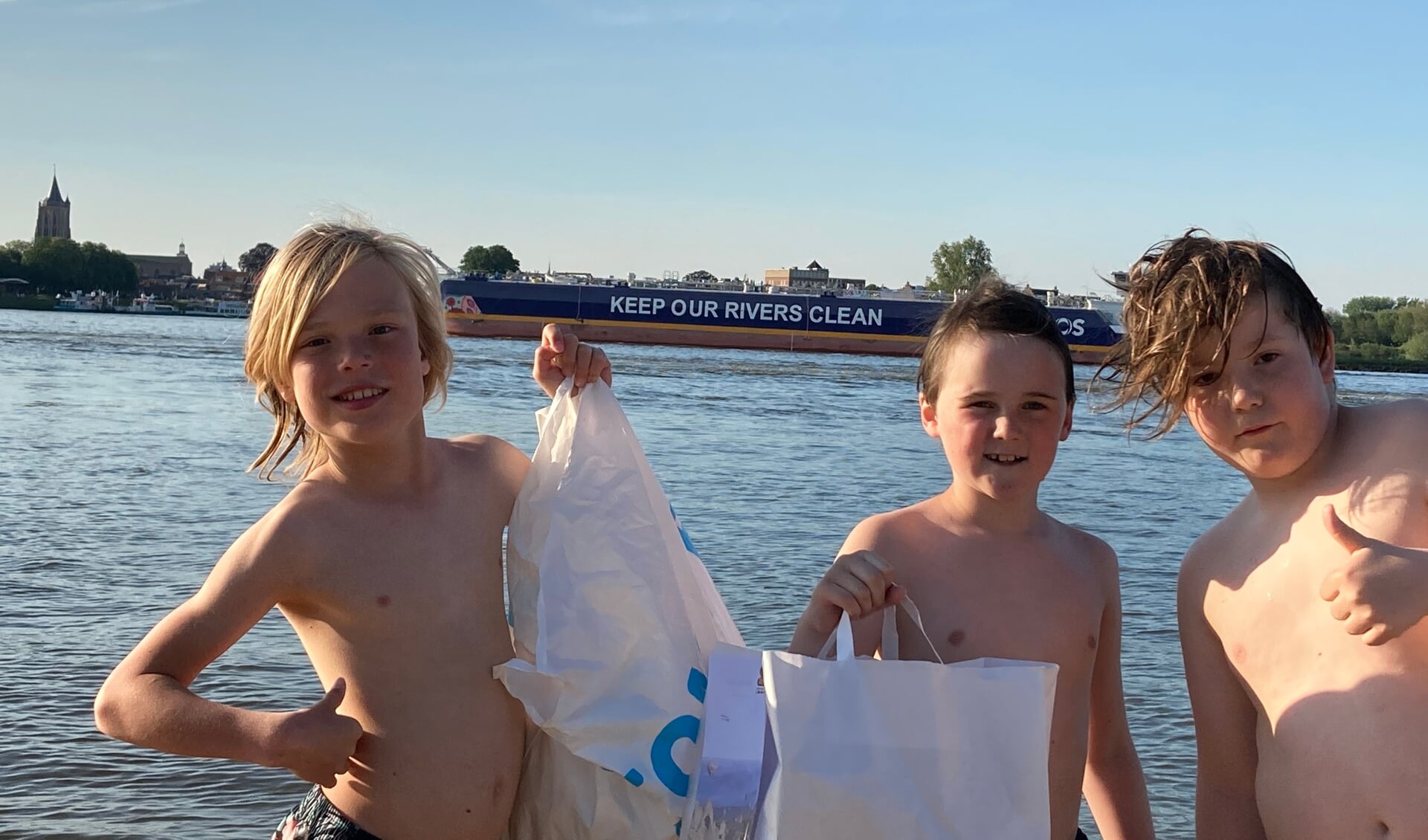 Maas, Dex en Mika met een stranddag afval en op de achtergrond het schip met toepasselijke boodschap.