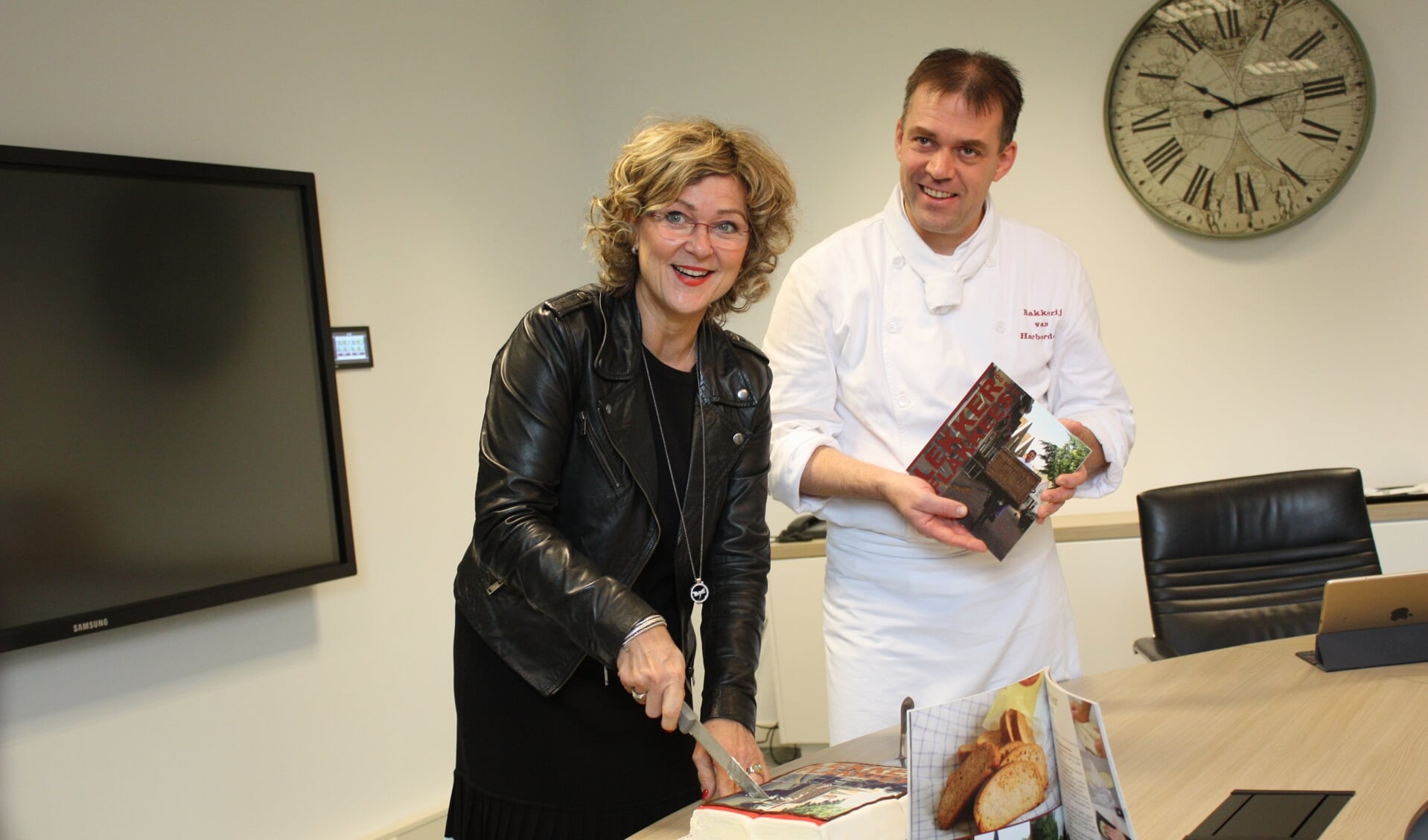 De burgemeester snijdt de taart aan met de cover van het nieuwe receptenboek. 