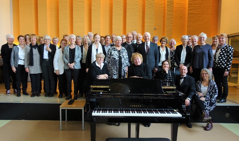 Achter de piano dirigent Carole Kroese (rechts) en stemcoach Elise Blesgraaf met het voltallige koor.