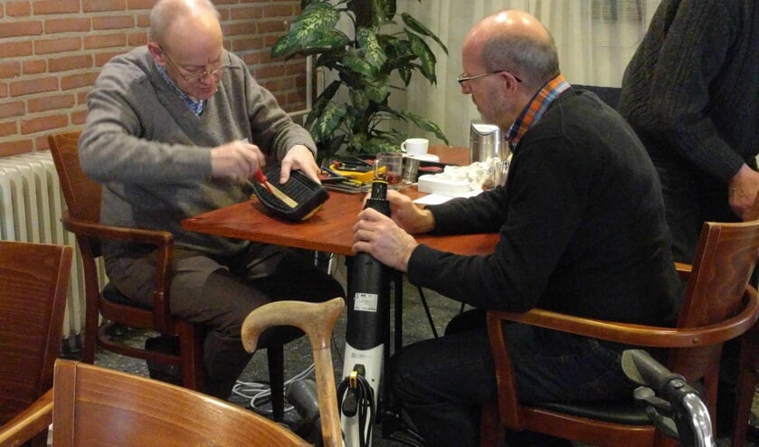 Een vrijwillige reparateur is bezig met een kapot apparaat, de eigenaar kijkt toe.