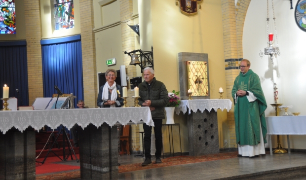 Nel van Schagen poseert trots met haar onderscheiding, naast haar staat haar echtgenoot. Pastoor Tilma kijkt toe.