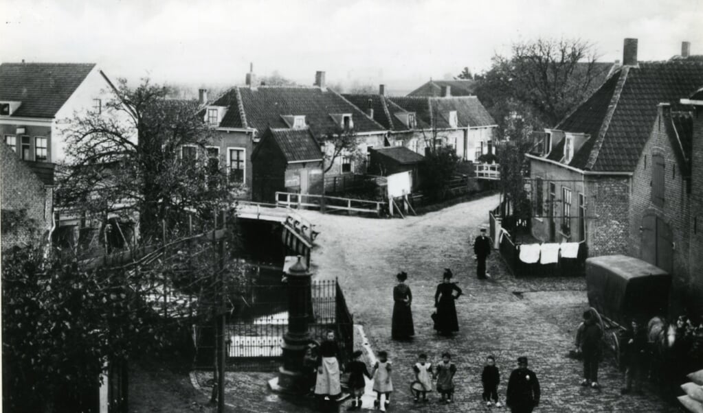 Het centrum van het dorp Kethel omstreeks 1905, gezien vanuit een woning op de hoek van de Dorpsstraat, Noordeinde en Schiedamseweg, in de richting van de Kerklaan, de weg naar links bij het witte hek. Op de voorgrond staan kinderen bij de 'Verspeekpomp'. Rechts de paardenstal van de Vergulde Valk. 