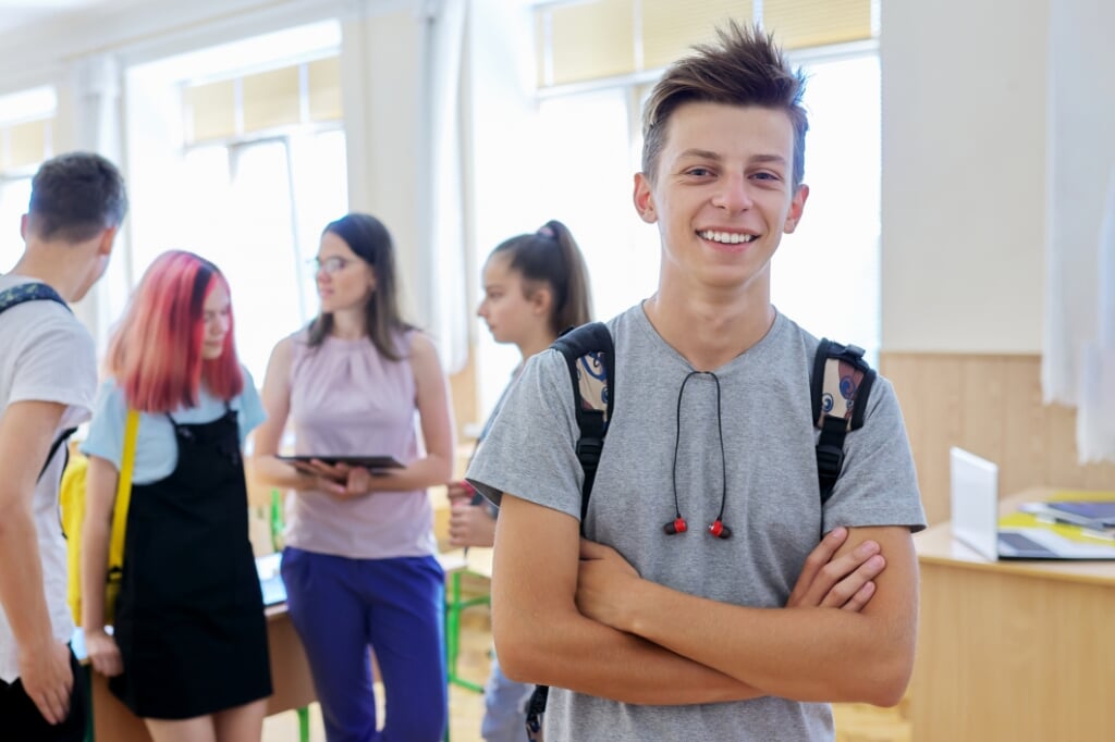 Portrait of smiling teenage boy in class on break. Background group of talking teenage classmates and teacher. School, college, study, youth concept (Portrait of smiling teenage boy in class on break. Background group of talking teenage classmates and
