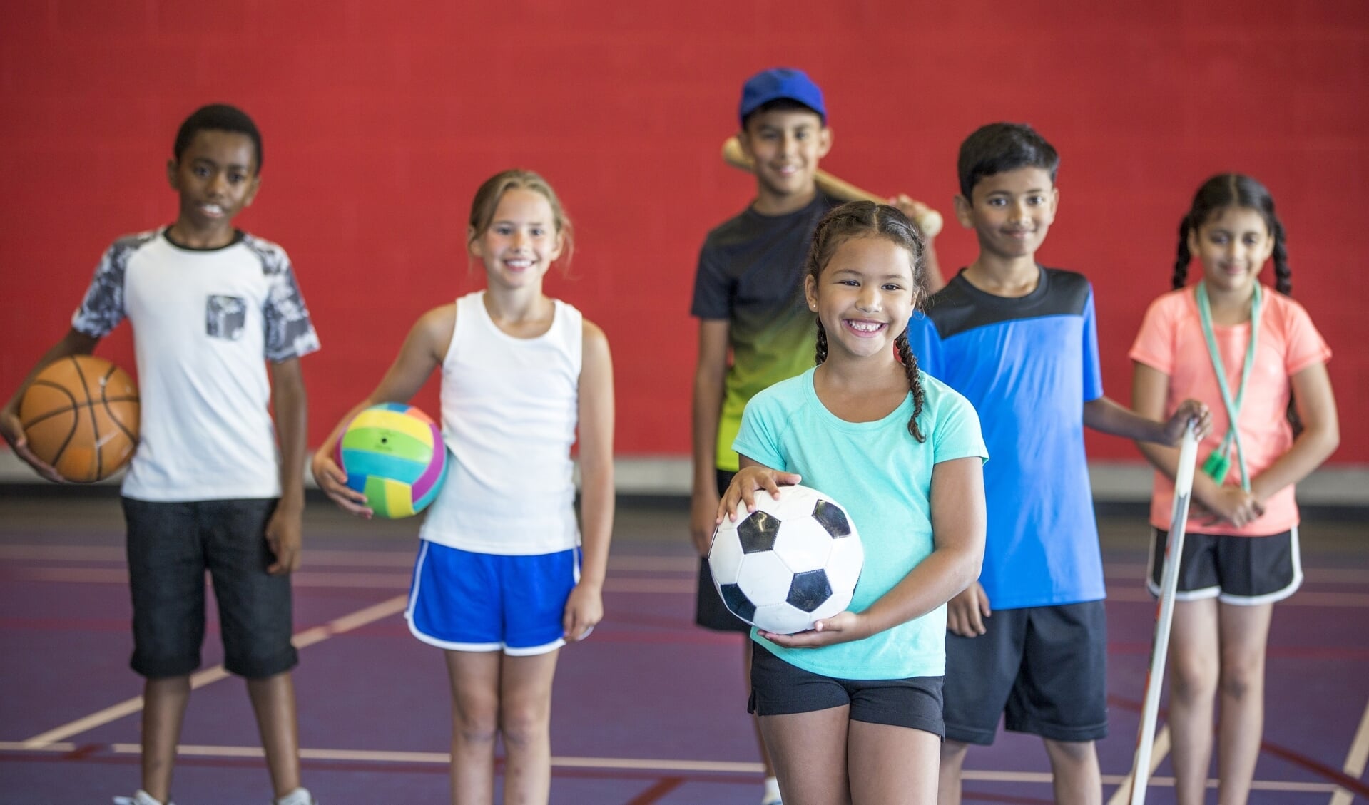 A multi-ethnic group of elementary age children  are playing various sports together in the gym. They are holding a basketball, soccerball, volleyball, and a hockey stick.