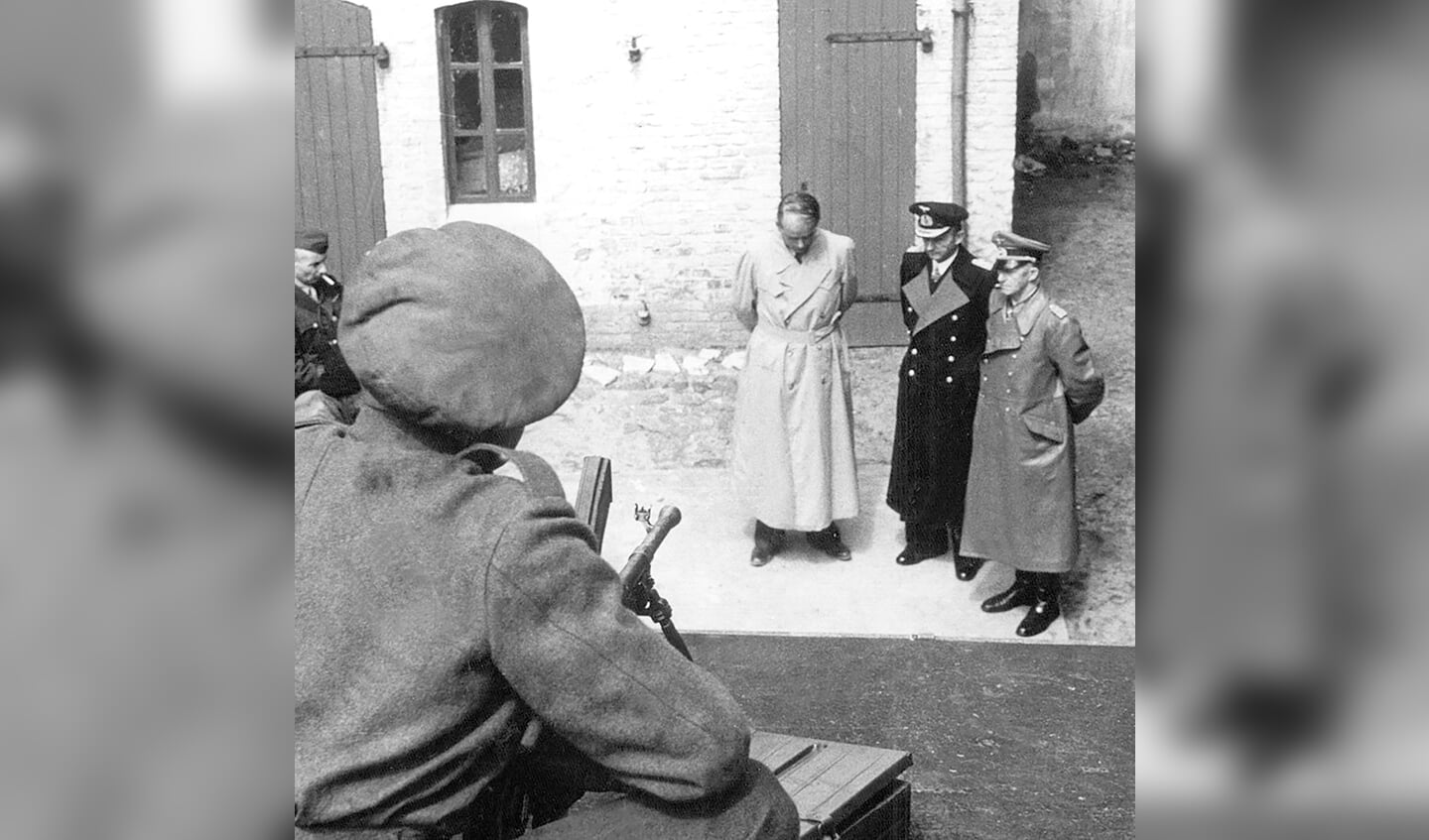 En britisk soldat overvåger fra et tag bag politigården i Flensborg arrestationen af tre nazi-spidser. Fra venstre er det rustningsminister Albert Speer, den sidste statschef i Det tredje Rige, Karl Dönitz, og hærchef Alfred Jodl.