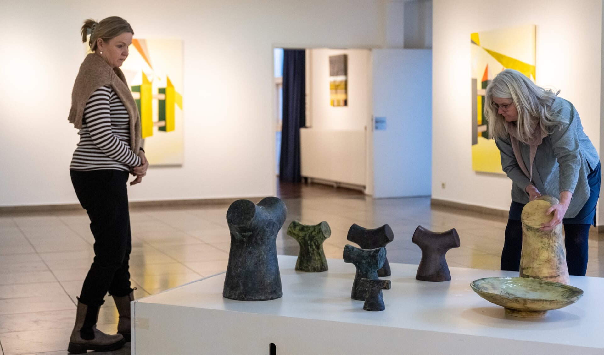Helle Bovbjerg (t.h.) stiller nogle af sine keramiske arbejder op i udstillingslokale på Flensborg Biblioteket, mens Annette Gerlif ser til. Hendes malerier kan ses i baggrunden. Foto: