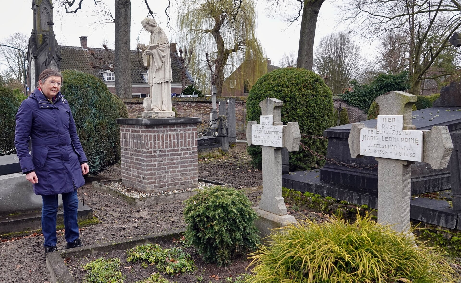Ricky van Oorschot bij twee graven van JMJ-zusters op het oude kerkhof van Gemonde. Op de achtergrond het beeld van Sint-Odulphus dat het ooit de gevel van het vroegere klooster sierde.