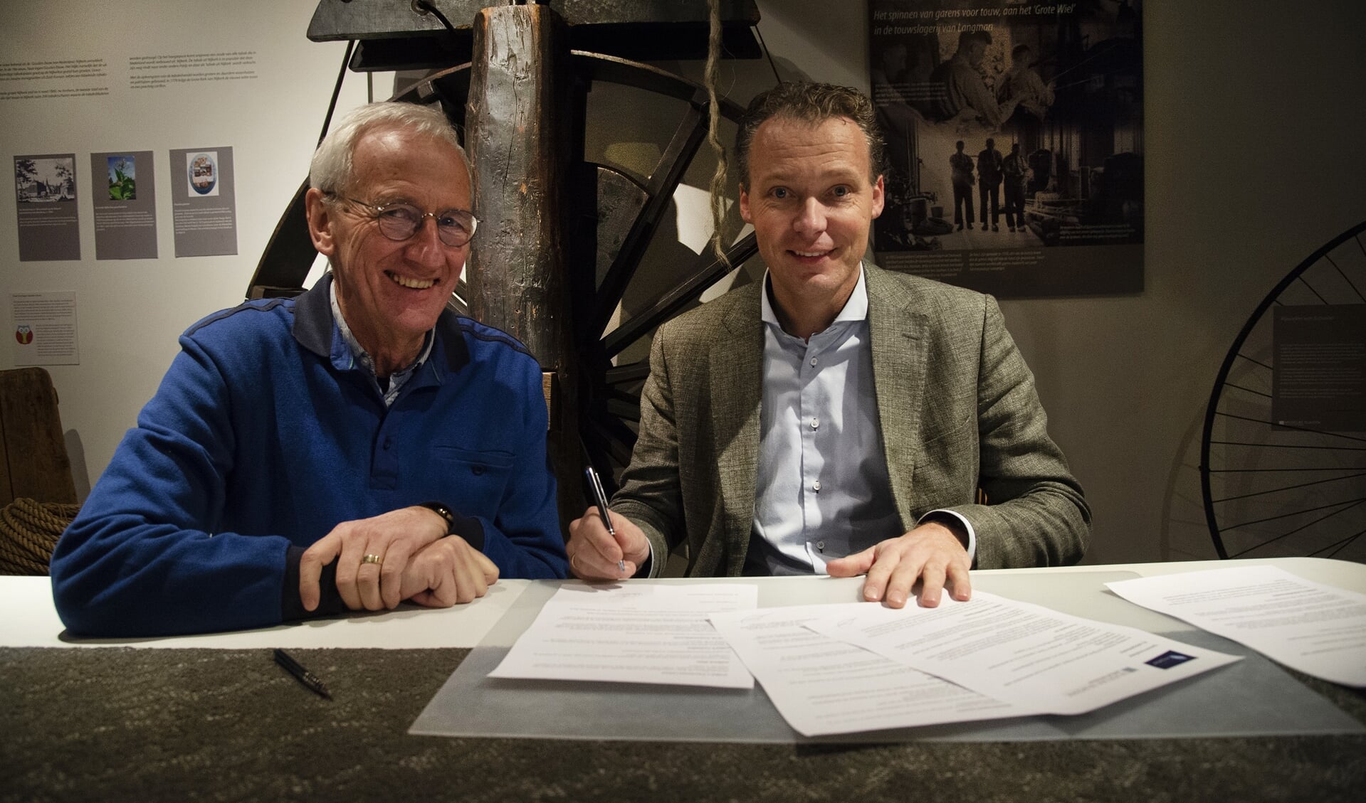 Jan Langman, directeur Touwfabriek Langman (rechts en Jan Cozijnsen, voorzitter Stichting Oud Nijkerk (links), ondertekenen het vernieuwde sponsorcontract. Op de achtergrond het grote wiel van de touwslagerij.