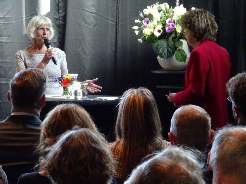 Dagvoorzitter Tineke van Booi interviewt Femke van Trier de Koningin luistert aandachtig.