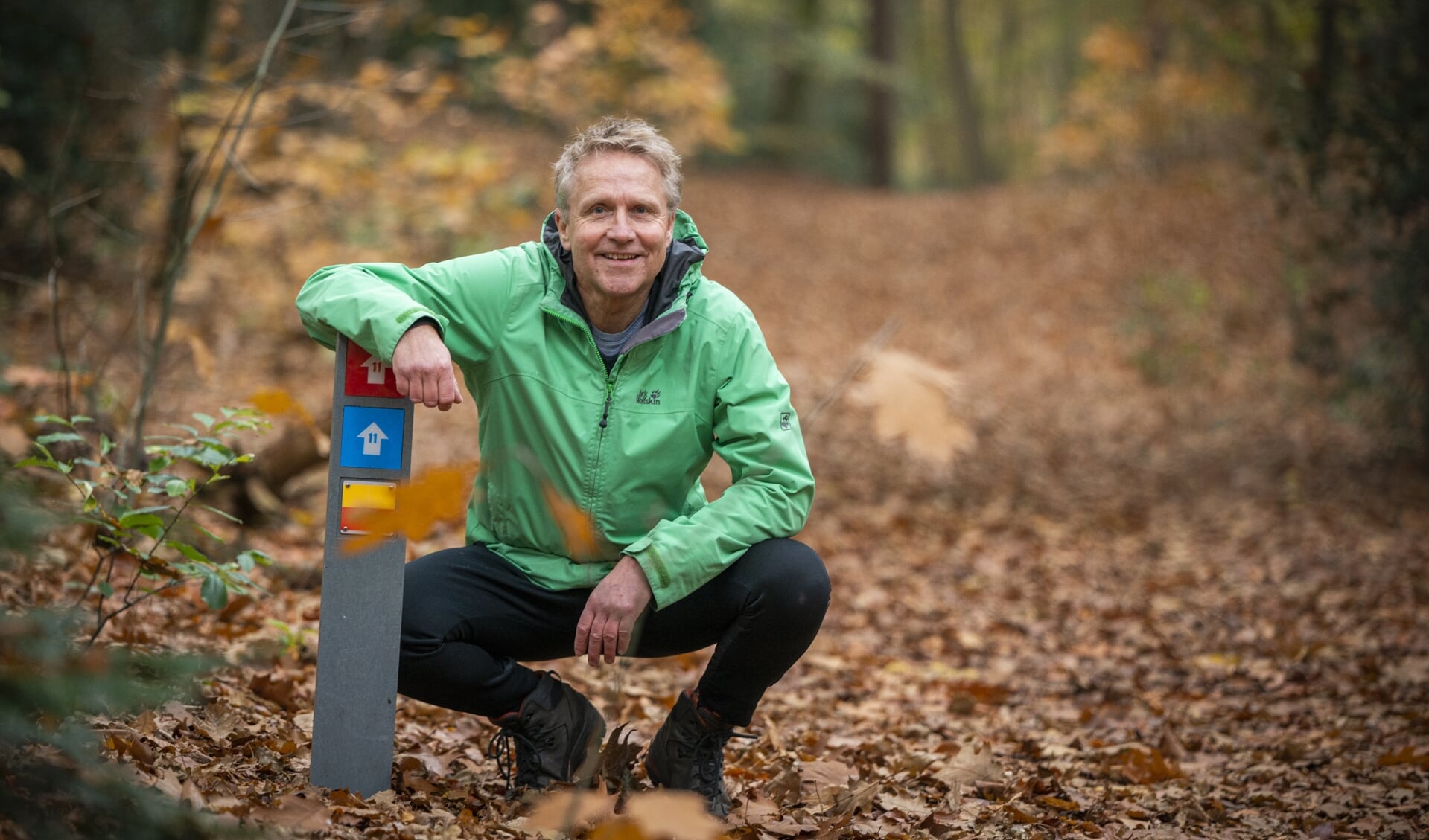 Henk van Geerenstein in 'zijn' Luntersche Buurtbosch bij de geel/rode markering van het Veluwe Zwerfpad en twee bordjes van het wandelnetwerk Goudsberg-Middelpunt van Nederland in Lunteren.