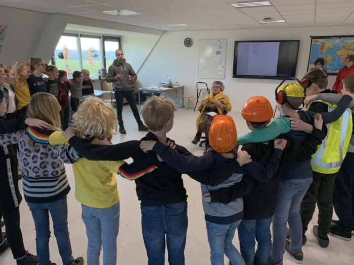 Groep 8 voert alvast een (mini-)musical op, speciaal voor juf Wilma. Thema: wat doet juf Wilma allemaal? Onder andere een verbouwing in goede banen leiden, vandaar de helmen. Wilma geniet in het midden van het lokaal.