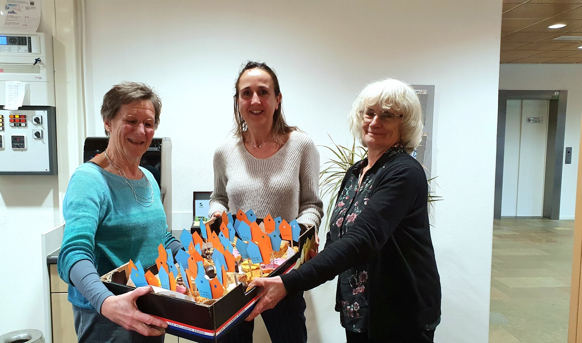 Nora Bakker (li) en Margaret Paige (re) bieden Mariëlle Versluis gebak aan als dank voor het gebruik van de keuken en kantine bij Kind & Co, 
