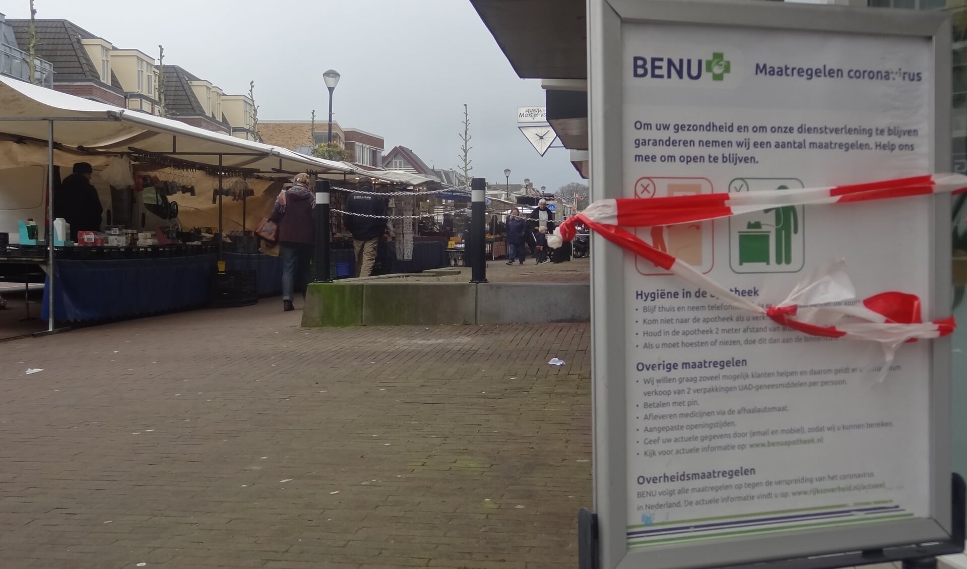 Een mededelingsbord met voorzorgsmaatregelen (en lint) bij de Benu Aportheek Soestdijk. Op de achtergrond draait de markt gewoon door.