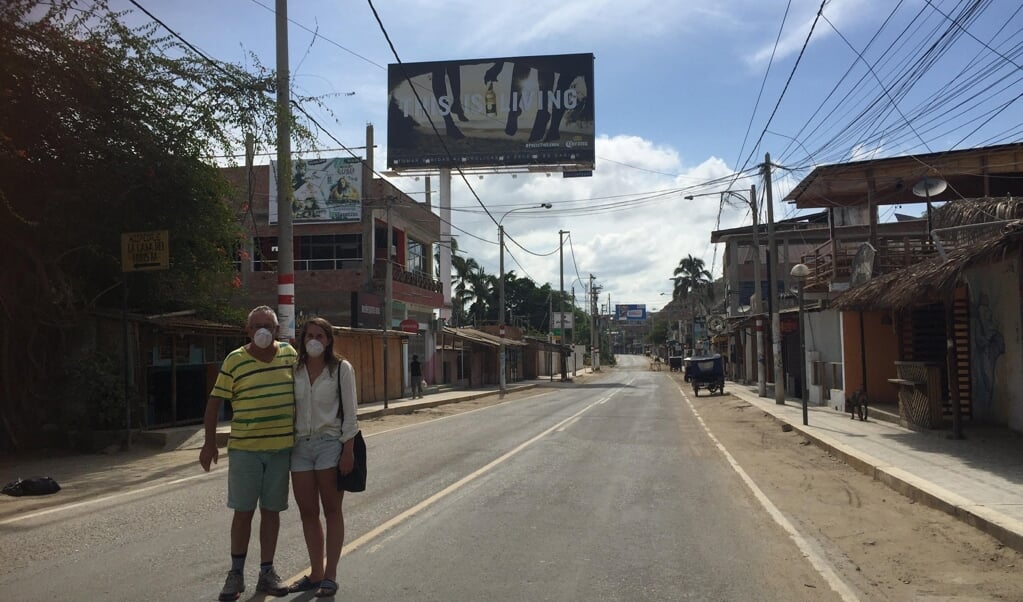 Herman Posthuma en dochter Dewi met mondkapje voor in een nagenoeg verlaten straat in Noord-Peru. Boven het asfalt een reclamebord: 'This is living, corona'.