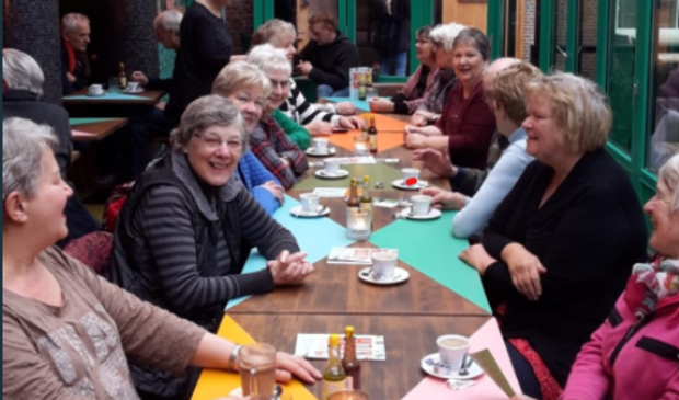 Mensen zitten aan lange tafel in cafe aan kopje koffie.