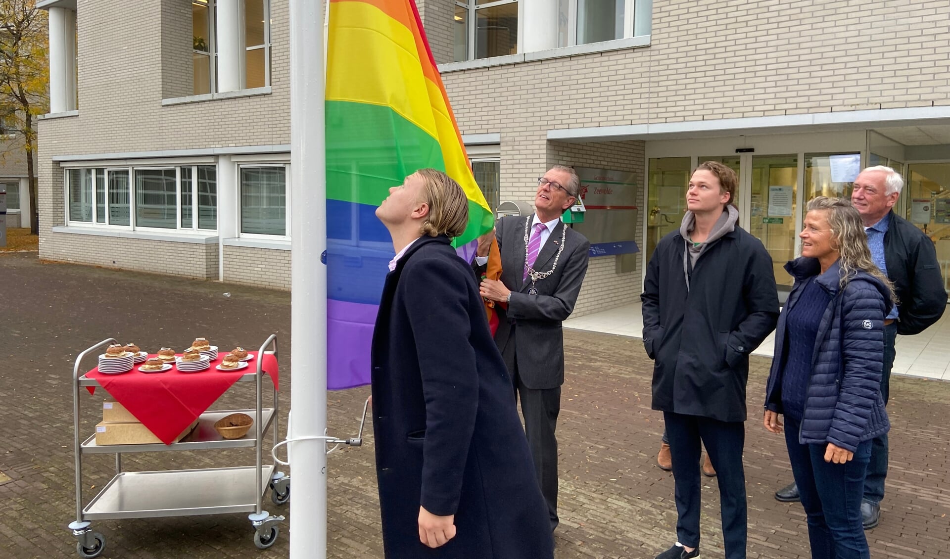 Maarten, de zoon van Gerrit van der Meer, hijst de regenboogvlag. Burgemeester Gorter assisteert. De gebakjes (optimistjes!) staan al klaar