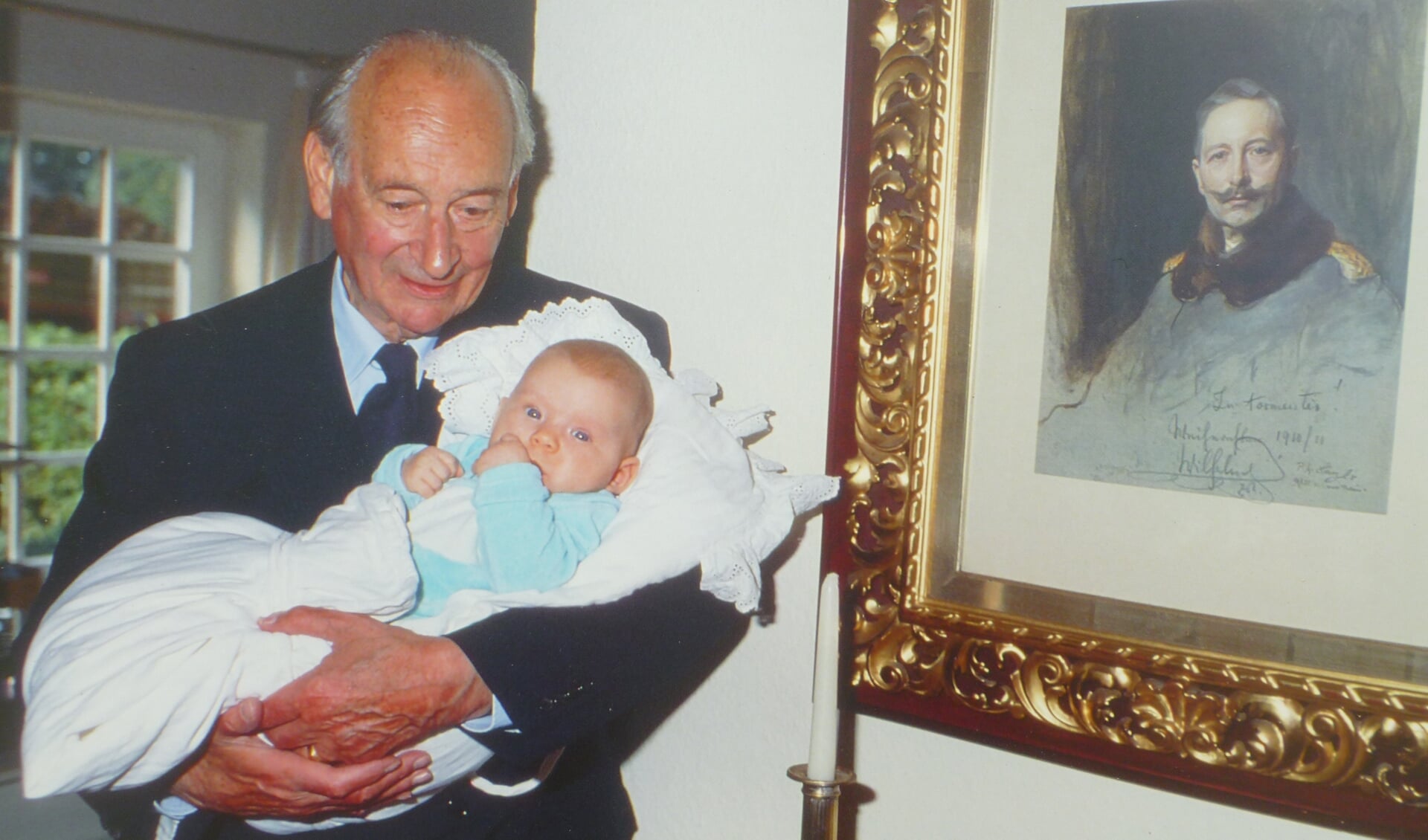 Prins Louis Ferdinand van Pruisen met in zijn armen één van zijn kleinkinderen. Rechts op een schilderij is zijn grootvader Keizer Wilhelm II van Duitsland afgebeeld. 