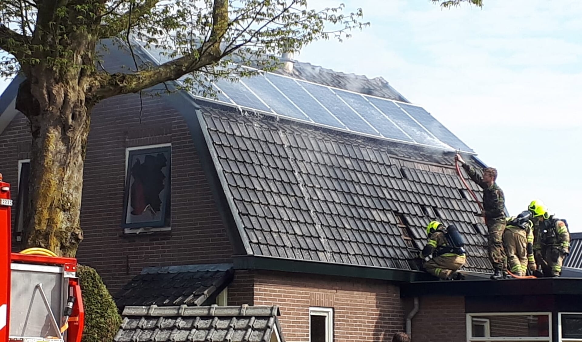 Terwijl net gearriveerde brandweerlieden dakpannen verwijderen, houdt een militair met een tuinslang het dak nat.