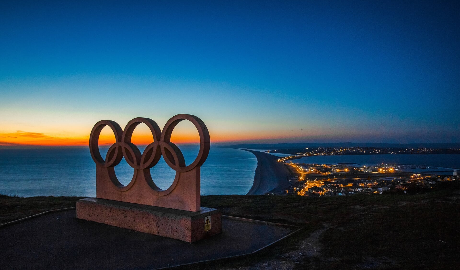 Een beeld van het logo van de Olympische Spelen met op de achtergrond de zonsondergang en uitzicht op een kustlijn en stad