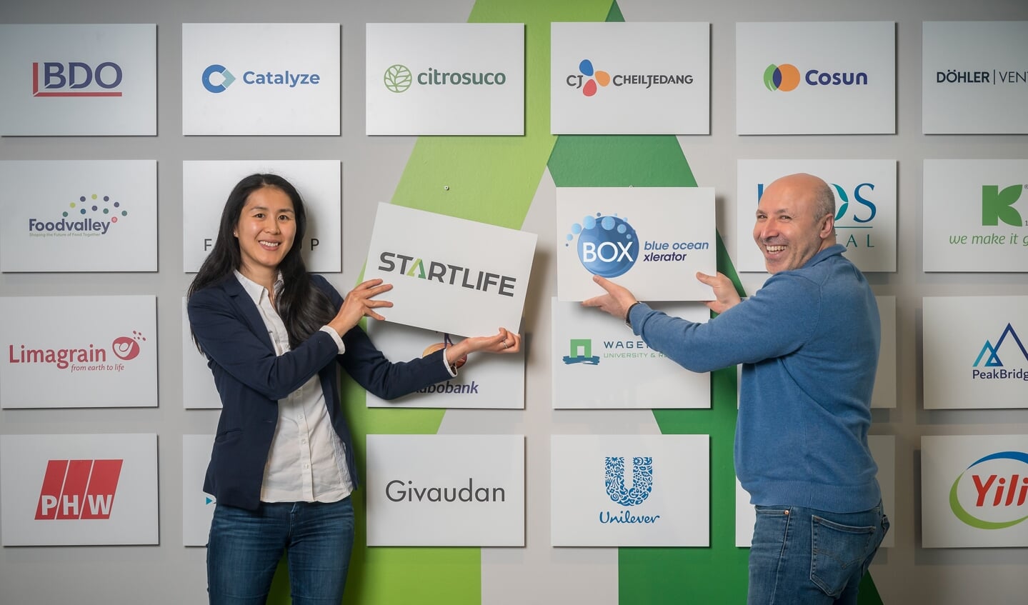 Lin Zhu (Investor Relations Manager, StartLife) and Rasit Görgülü (Partner, BOX) hangen logo's op aan StartLife's partnermuur