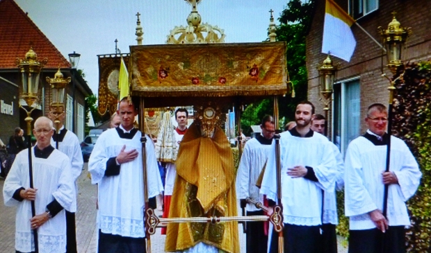 Pater Carlo de Beer, die tijdens de Sacramentsprocessie de Monstrans met de Hostie in zijn Handen draagt, loopt onder het baldakijn, dat door vier baldakijndragers wordt vastgehouden.