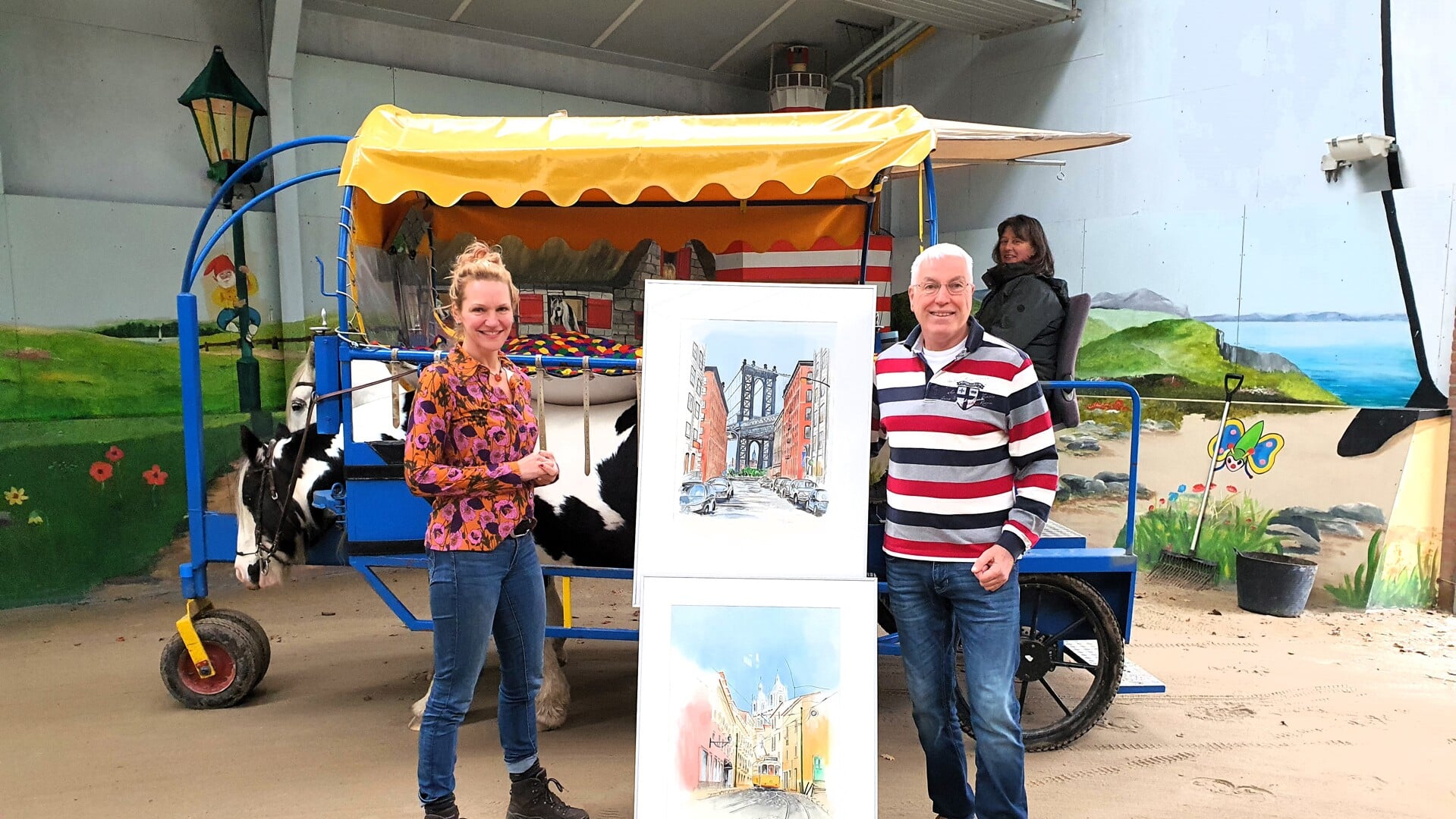 Esther Lucassen en Theo Janssen poseren bij een huifbed en twee van Janssens schilderijen die op 18 november tijdens de expositie ook te zien zullen zijn.