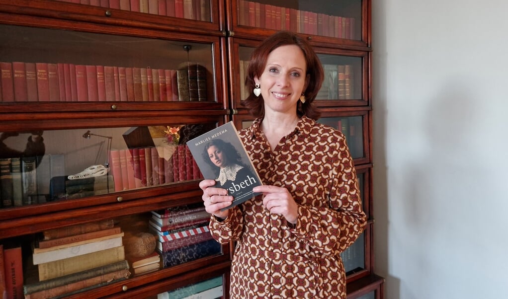 Marlies bij een boekenkast vol historische verhalen en met haar eigen roman in de hand