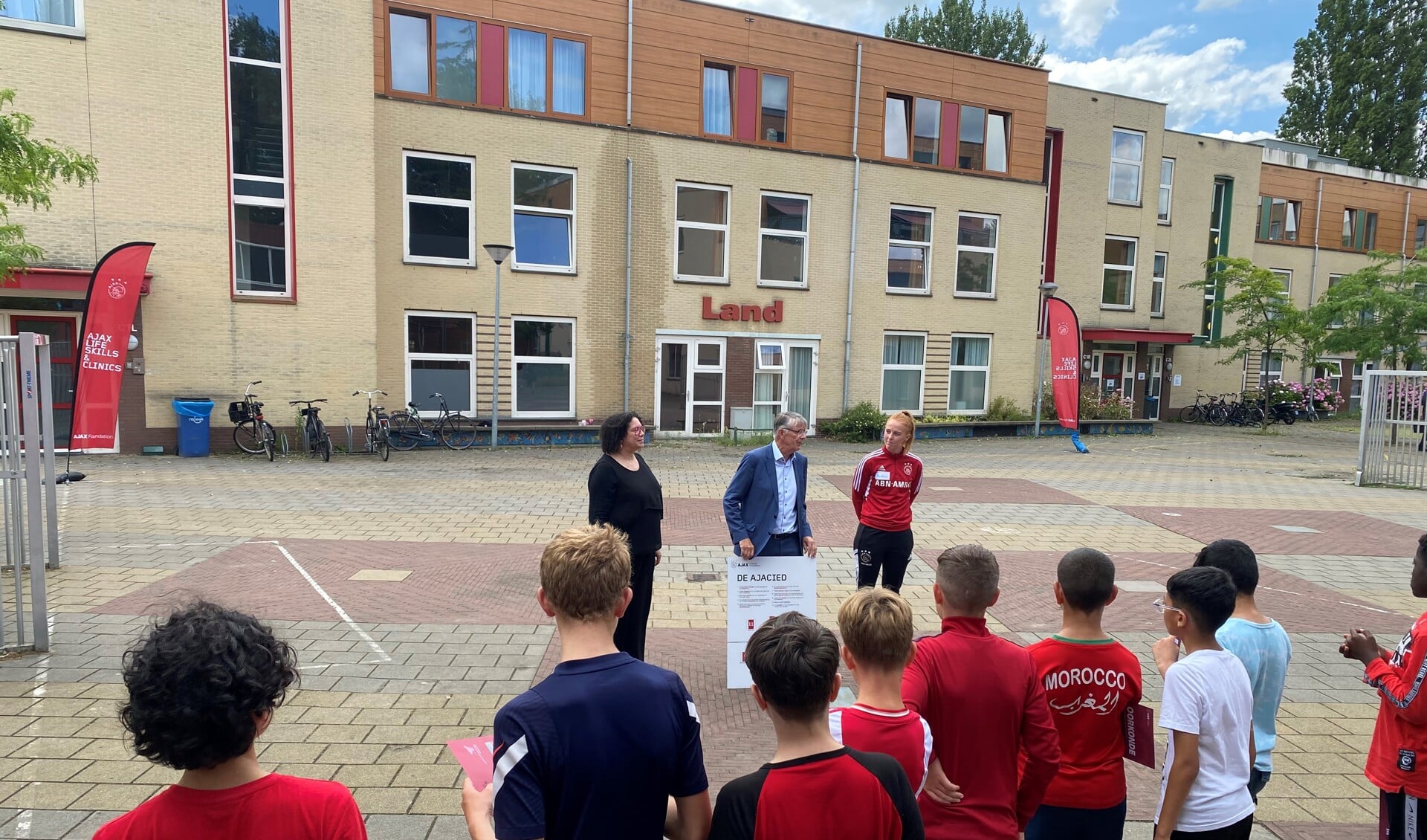 Wethouder Frequin (Sport, VVD) met het ‘Bord met Kernwaarden’ voor de deelnemers en directie (links van Frequin) van De Pionier. Rechts in Ajax tenue: Amber Wuring (programmabegeleding van de Ajax Foundation).