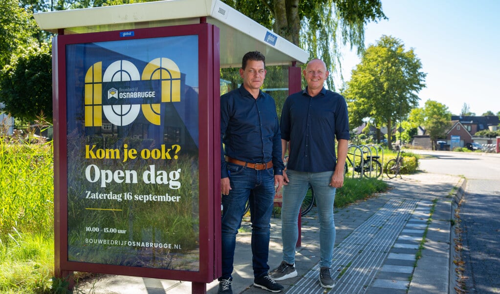 Directeuren Henk ten Broek en Marco van den Essenburg van Bouwbedrijf Osnabrugge bij de bushalte voor hun bedrijf, die opgesierd is met de uitnodiging voor de open dag.