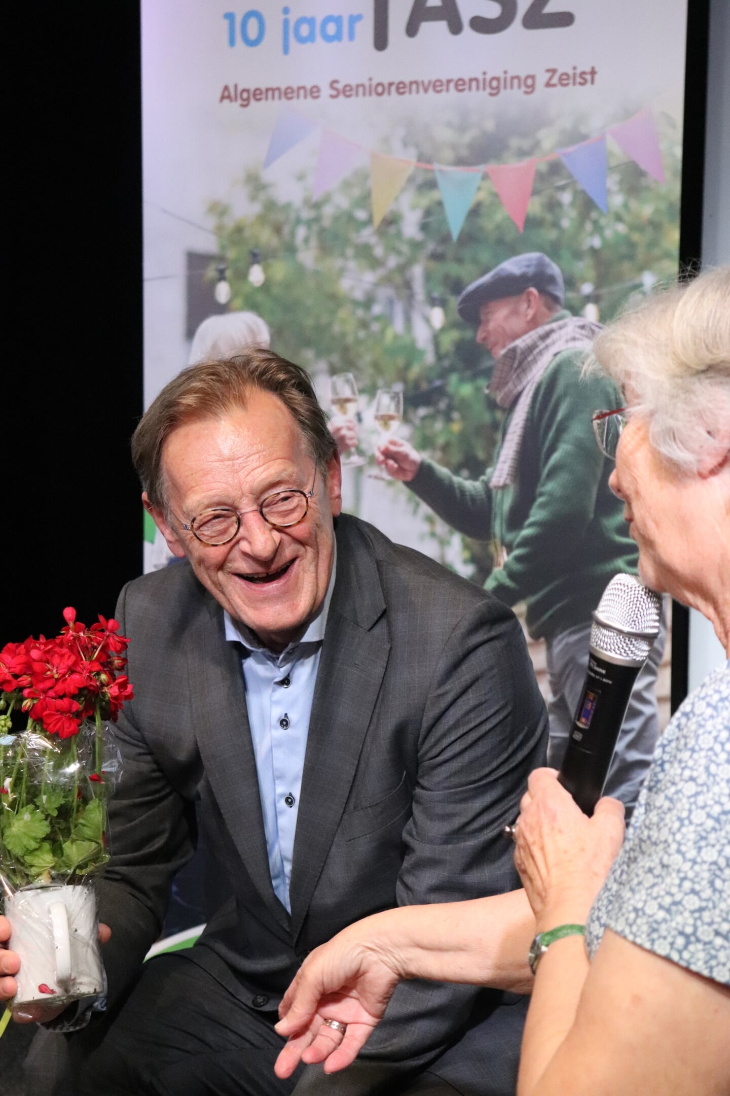 Koos Janssen krijgt een geranium van Lia van Dijk, voorzitter van de ASZ, maar hij mag er niet achter gaan zitten.