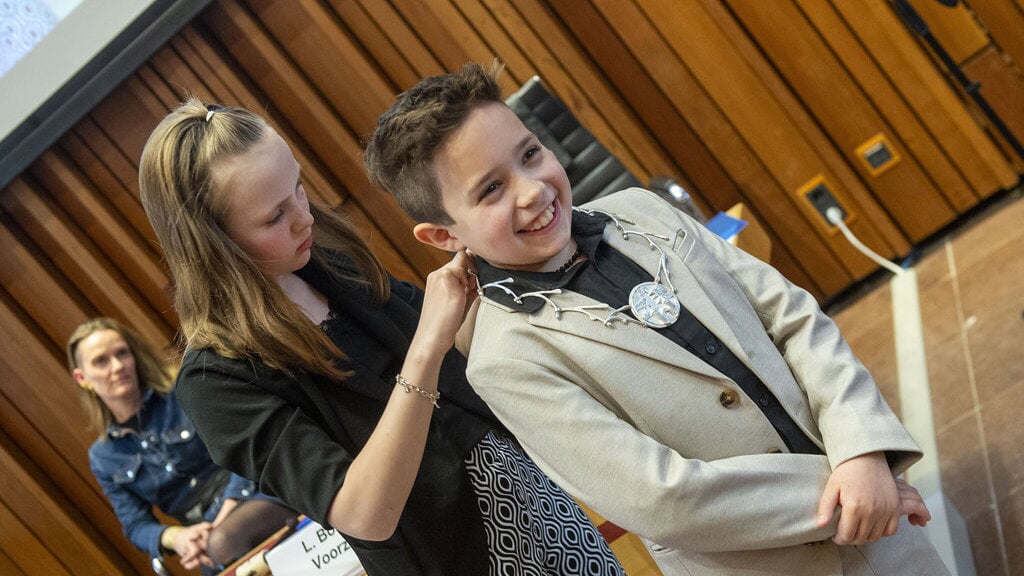 Aiden krijgt de ambtsketen van kinderburgemeester omgehangen door de oud-kinderburgemeester Lynn Verhoeven.