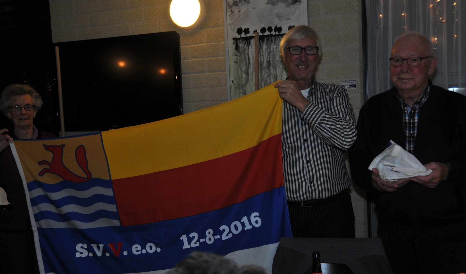 Voorzitter Jos Hommel en Anna Stoutjesdijk (zij is het langst lid) ontvouwen de nieuwe vlag. Rechts houdt bestuurslid Gerrit de Gast de oude Anbo-vlag vast.