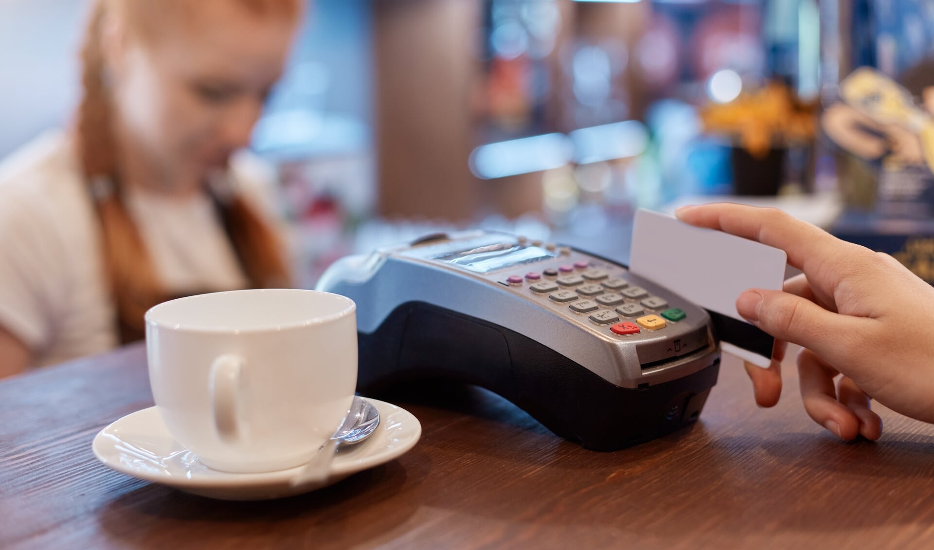 Customer paying for cup of coffee with card, using terminal for paying, white cup of coffee and red haired barista on background, faceless person uses device and card.